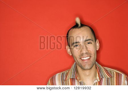 Caucasian man with mohawk smiling against red background.