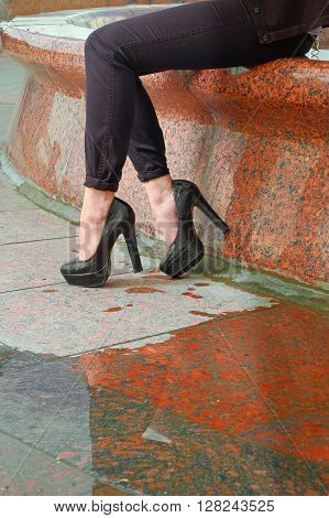 Closeup of young woman legs on red granite cobblestone street with puddle and reflection. Beautiful female legs in fashion shoes with high heels on granite pavement vertical shot. Instagram effect.
