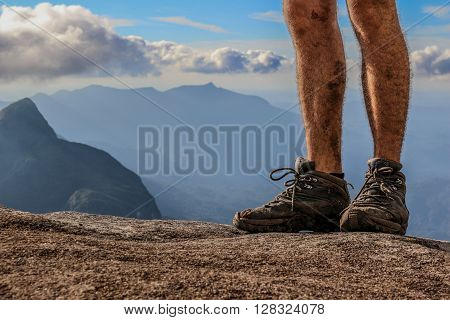 Man hiker with dirty legs and hiking boots on mountain peak rock with a valley at background