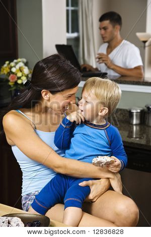 Donna caucasica con figlio bambino in cucina con il padre sul portatile in background.