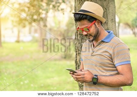 Happy indian guy looking his mobile phone and smiling - Young asian man texting online in park outdoor - New technology trends addiction concept - Warm filter with focus on face