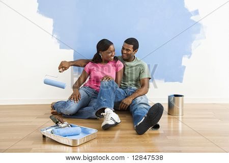 Africká americká pár relaxační společně vedle napůl malované zdi a malířské potřeby.