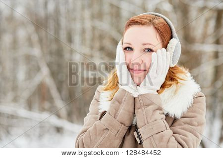 Humeral portrait of red-haired girl in earmuffs, clasping face with her hands in white knitted gloves. 