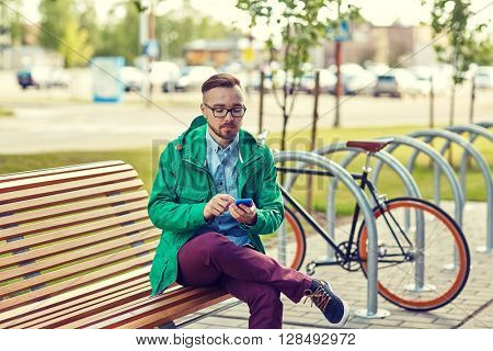 people, style, technology and lifestyle - happy young hipster man with smartphone and fixie bike sitting on bench in city