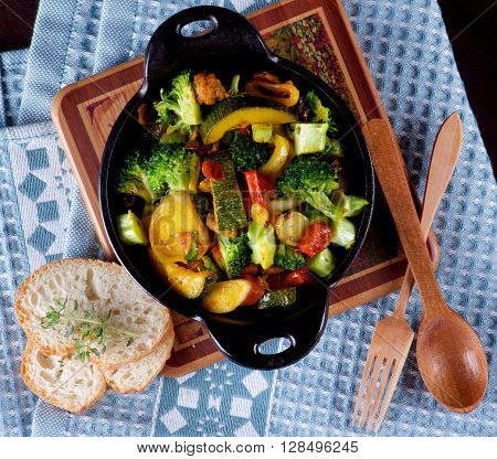 Homemade Colorful Vegetables Ragout with Zucchini Carrots Broccoli Leek and Red Bell Pepper in Black Iron Stewpot with Wooden Spoon and Fork and Bread closeup on Blue Napkin. Top View
