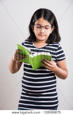 indian small girl with eye glasses standing and reading a book, asian little girl holding a book and reading over white background, cute indian girl wearing spectacles and reading book while standing