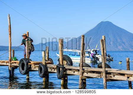 Lake Atitlan Guatemala - April 4 2016: Local man dressed in traditional Mayan pants pulls in boat from its mooring in early morning light with San Pedro volcano behind on Lake Atitlan, Guatemala.