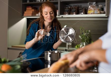 Happy young couple baking and cooking meal at kitchen. Husband helping wife in preparing lunch. Woman adding pepper in pan for taste. Woman pouring salt in the pot.