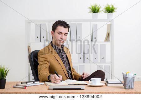 Caucasian businessman in brown jacket sitting at office desk and reflecting something in journal