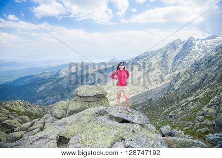 brunette woman with red sweater brown short trousers and sunglasses posing on top rock looking over peaks mountain and valley in Gredos mountain Avila Spain Europe