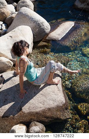 brunette woman green shirt grey trousers barefoot sitting on rock touching water feeling temperature in pond of river lake or lagoon in summer Gredos Avila Spain Europe