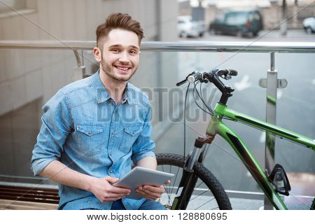 Modern generation.  Cheerful positive smiling young man sitting on the bench and holding tablet while expressing joy
