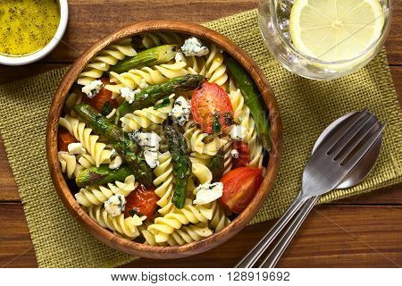 Baked green asparagus cherry tomato blue cheese and rotini pasta salad served in wooden bowl photographed overhead on dark wood with natural light
