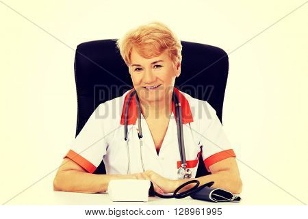 Smile elderly female doctor or nurse sitting behind the desk with bloog preasure gauge