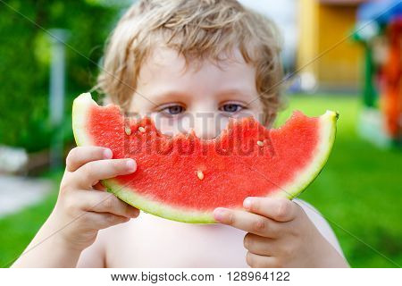 Adorable little toddler boy with blond hairs eating watermelon in summer garden. Kid tasting healthy snack. Selective focus on watermelon