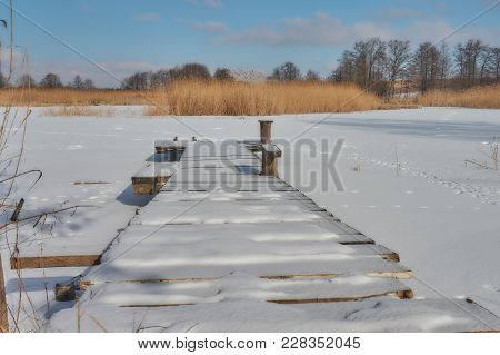 The Frozen River Bank, Snow Covered Footbridge, Trees, Rushes, Fallen Fields And Blue Sky With White