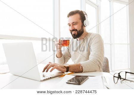European joyous man 30s with short brown hair drinking tea and listening to music via wireless headphones while using notebook