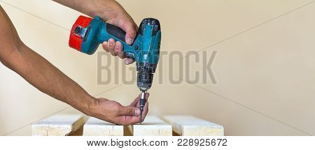 Hand Of A Worker Screws A Screw In A Wooden Board With A Cordless Screwdriver. Man Carpenter At Hand