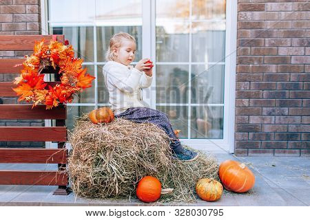 Adorable Smiling Caucasian Baby Toddler In White Knittes Jacket Sitting On The Haystack With Pumpkin