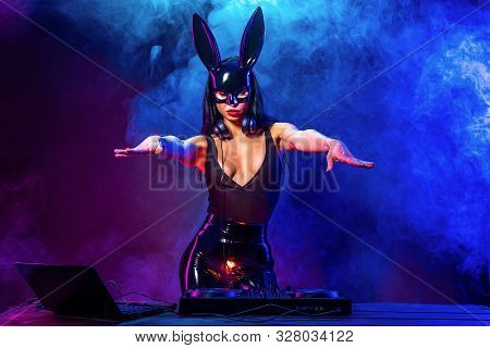 Young Sexy Woman Dj Playing Music In Mask. Headphones And Dj Mixer On Table. Smoke On Background