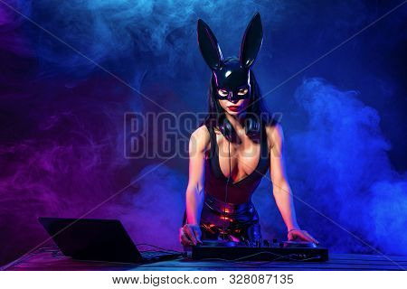 Young Sexy Woman Dj Playing Music In Mask. Headphones And Dj Mixer On Table. Smoke On Background