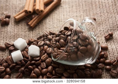 Scattered Coffee Beans And Sugar From A Glass Cup On Sackcloth, With Cinnamon Sticks Behind.