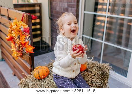 Adorable Blonde Baby Toddler In White Knittes Jacket Sitting On The Haystack With Pumpkins At Porch,