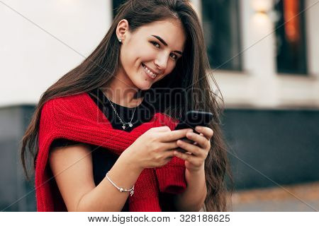 Portrait Of Smiling Positive Young Woman Sitting Outdoors Texting On Mobile Phone. Brunette Female W