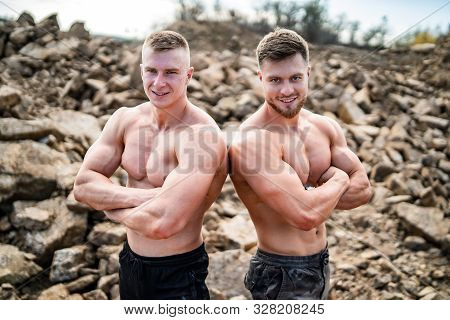 Two Fitness Strongmen Standing Back To Back With Crossed Hands In A Rocky Background. Bodybuilders C