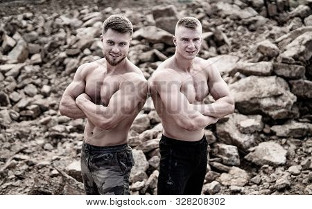 Two Fitness Strongmen Standing Back To Back With Crossed Hands In A Rocky Background. Bodybuilders C