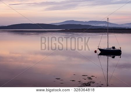 Sailing Boat Reflecting In A Calm Loch As The Sun Sets Casting Pink Tones Across The Water On Partia