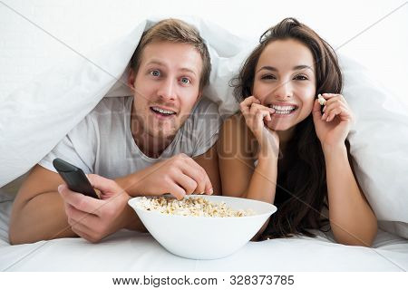 Young Couple Handsome Man And Beautiful Woman Watching Tv In Bed Eating Pop Corn Looking Happy