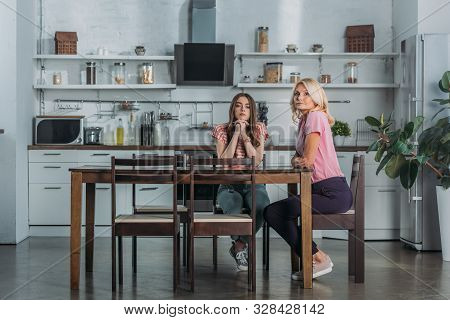 Attractive Mature Woman With Young Girl Sitting At Kitchen Table And Looking At Camera