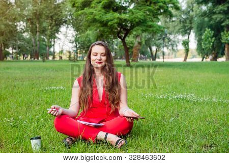 Young Businesswoman In Red Suit Sitting In Lotus Position On Green Grass In Park And Holding Her Sma