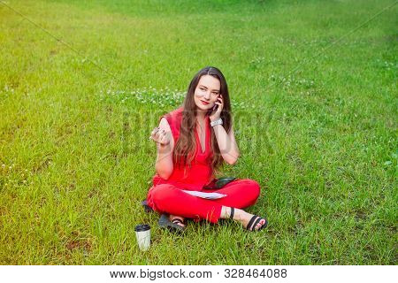 Young Business Woman Plus Size In Red Suit Speaking Her Phone And Seating On The Background Of Green