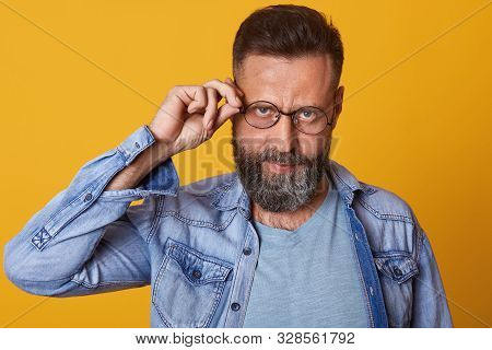 Photo Of Middleaged Bearded Man Keeps Hand On Rim Of Spectacles, Wearing Casual Clothes, Poses Again