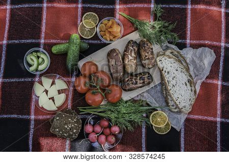 Picnic Red Plaid On Which Lies A Picnic Food, Barbecue, Sausages On The Fire, Fresh Vegetables, Brea