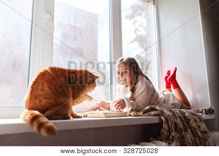 Little Girl In Warm Terry Robe Lying On Window Sill And Reading A Book. Winter Weekend With Cat At H
