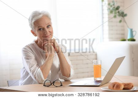 Mature Woman With Laptop Sitting At Table In Kitchen. Smart Aging
