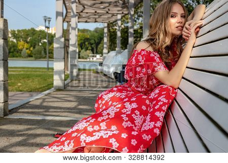 Beautiful Young Girl, Model, Brown-haired Woman With Long Hair In A Red Dress Sits On A Bench And Po