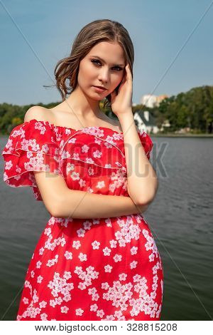Portrait Of A Beautiful Young Brown-haired Girl With Long Hair In A Red Dress On A Lake Background.