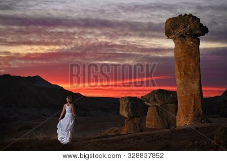 Romantic Image. Blond Woman In White Dress By The Giant Rock Toadstools Watching Sunset In Rock Dese