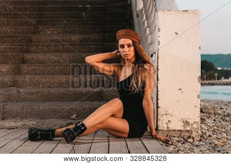 Sexy Blonde In A Black Dress And Hat Posing Sitting On The Steps. In The Distance You Can See The Se
