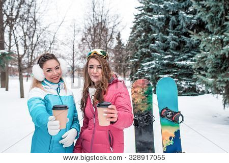 Two Girls Girlfriends Winter Park Resort Hold Out Cups Hot Coffee Tea, Offer And Treat Them Share. E