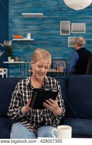 Pensioner Lady Browsing On Internet Sitting On Couch. Elderly Woman Using Moder Technoloy Tablet Pc 