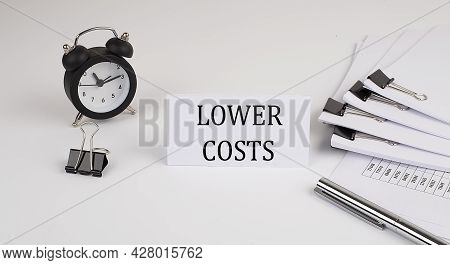Card With Text Lower Costs On A White Background, Near Office Supplies And Alarm Clock. Business