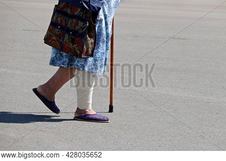 Old Woman With Bandaged Leg Walking With A Cane Down The Street. Concept Of Injury And Wound, Elderl