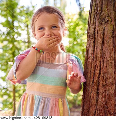 Smiling Young Girl Playing Hide And Seek Behind Tree In Garden