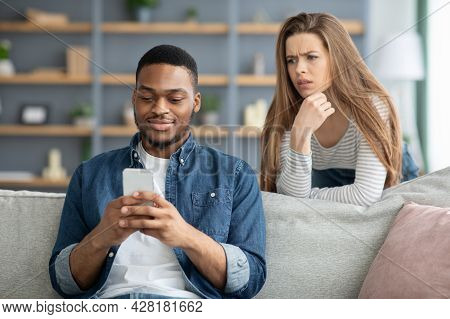 Mistrust. Attentive Lady Looking At Black Boyfriend While He Texting On Smarthone