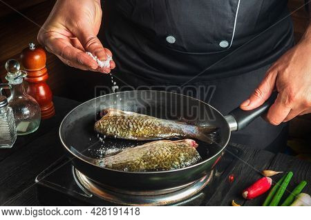 Professional Chef Prepares Fresh Fish In Pan Sprinkling Salt With The Ingredients. Preparing To Cook